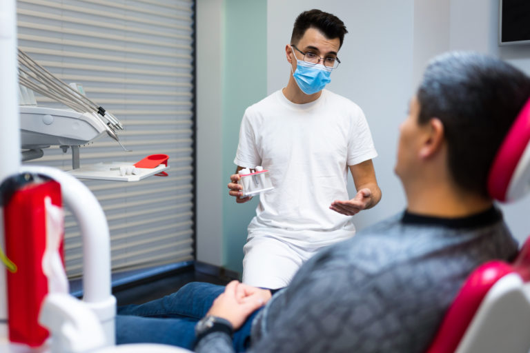 Dental Surgeon With Dental Crown In Hands Model Talks To Patient About Prosthetics Man At The Dentist S Appointment